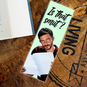 May include: A bookmark with a light green background featuring a man with glasses and the text "Is that smut?". The man is holding a book. The bookmark is on a wooden surface.