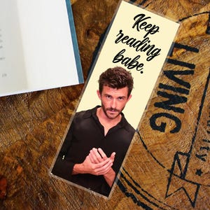 May include: A bookmark featuring a man in a black shirt, with the words "Keep reading babe." The bookmark is placed on a wooden surface, beside an open book.