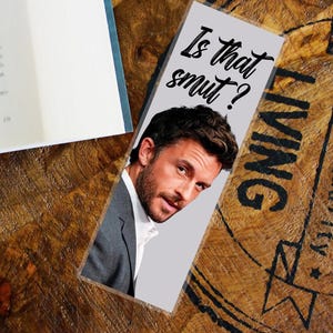 May include: A bookmark with the text "Is that smut?" in black script. The bookmark features a photo of a man in a gray suit and white shirt. The bookmark is on a wooden surface next to a book.