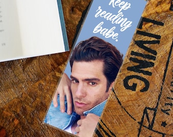 May include: A bookmark featuring a man in a denim jacket with the text "Keep reading babe." The bookmark is rectangular and has a light blue background. The bookmark is on a wooden surface.