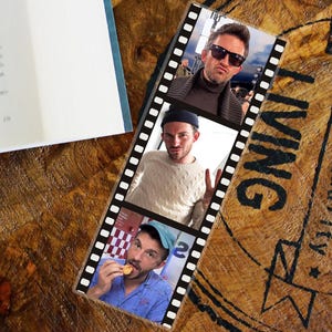May include: A bookmark with a film strip design, displaying three images of a man. The man is wearing sunglasses, a beanie, and a blue cap. The bookmark rests on a wooden surface, with the word "LIVING" visible.