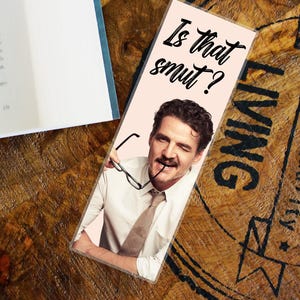 Peut inclure: Marque-page avec un fond rose clair représentant un homme faisant un clin d'œil et tenant des lunettes. Le texte "Is that smut?" est écrit en cursive noire. Le marque-page est sur une surface en bois à côté d'un livre.