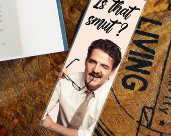 May include: A bookmark with a light pink background featuring a man winking and holding glasses. The text "Is that smut?" is written in black cursive. The bookmark is on a wooden surface next to a book.