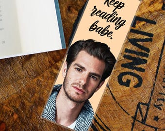 May include: A rectangular bookmark with a photo of a man and the text "Keep reading babe." The bookmark is peach-colored with black text and a photo of a man in a blue and white patterned shirt.