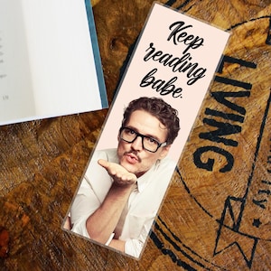 Può includere: Un segnalibro rettangolare con sfondo rosa chiaro raffigura un uomo con gli occhiali che manda un bacio. Il testo "Keep reading babe." è scritto in corsivo nero. Il segnalibro è su una superficie di legno accanto a un libro.