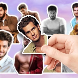 Könnte beinhalten: Eine Hand hält einen Aufkleber eines Mannes mit braunen Haaren und Bart. Der Aufkleber ist von anderen Aufklebern desselben Mannes in verschiedenen Posen und Outfits umgeben. Der Hintergrund ist ein weicher lila-weißer Farbverlauf.
