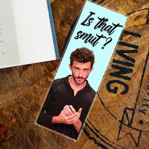 May include: A light blue bookmark with the text "Is that smut?" in black script. The bookmark features a photo of a man in a black shirt. The bookmark is on a wooden surface.
