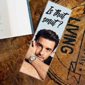 May include: A light blue bookmark featuring the text "Is that smut?" above a photograph of a man wearing a watch. The bookmark rests on a wooden surface.