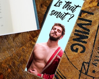 May include: A light blue bookmark with the text "Is that smut?" features a shirtless man with a red and gold patterned shawl. The bookmark is on a wooden surface next to an open book.