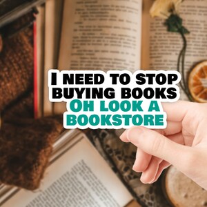 Op de afbeelding: Een sticker met de tekst "Ik moet stoppen met het kopen van boeken. Oh kijk, een boekwinkel." De sticker is wit met zwarte tekst en een turquoise rand. De sticker wordt vastgehouden door een hand.