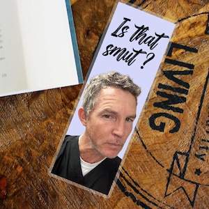Könnte beinhalten: Ein rechteckiger Lesezeichen mit hellviolettem Hintergrund zeigt das Gesicht eines Mannes und den Text "Is that smut?" in schwarzer Schrift. Das Lesezeichen liegt auf einer Holzoberfläche neben einem Buch.