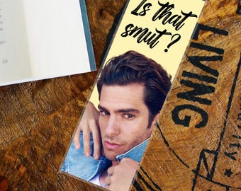 May include: A rectangular bookmark with a light yellow background features a photo of a man and the text "Is that smut?". The bookmark is on a wooden surface next to a book.