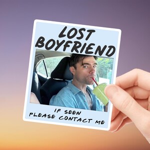 Op de afbeelding: Een lichtblauwe en witte sticker in polaroidstijl met de tekst "LOST BOYFRIEND" en "IF SEEN PLEASE CONTACT ME". De sticker toont een man die uit een groene beker drinkt. De sticker wordt door een hand vastgehouden.