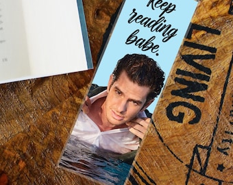 May include: A bookmark with a light blue background featuring a photo of a man and the text "Keep reading babe." The bookmark is on a wooden surface next to a book.