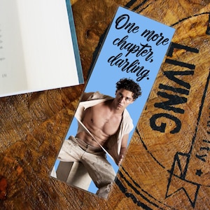 Könnte beinhalten: Ein rechteckiges Lesezeichen mit hellblauem Hintergrund und dem schwarzen Text One more chapter, darling. Unter dem Text befindet sich ein Foto einer Person mit lockigem dunklem Haar, die ein offenes beiges Hemd trägt.
