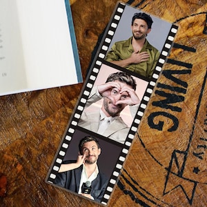 May include: A bookmark with a film strip design, displaying three images of a man. The man is shown in different poses and outfits. The bookmark is placed on a wooden surface, next to a book.