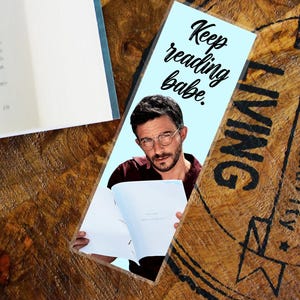 May include: A light blue bookmark featuring a man with glasses and the text "Keep reading babe." The bookmark is placed on a wooden surface next to an open book. The man is holding an open book.
