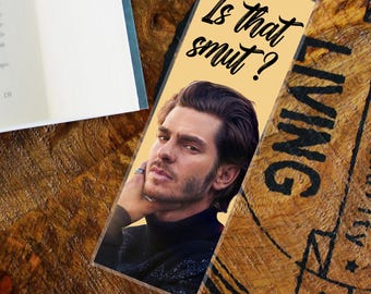 May include: A rectangular bookmark with the text "Is that smut?" above a portrait of a man. The bookmark has a yellow background and is placed on a wooden surface next to a book.