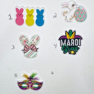 May include: A collection of six decorative charms. The charms include three Peeps, a white bunny with an Easter egg, a bunny with floral ears, a Mardi Gras design, and a purple masquerade mask with feathers.
