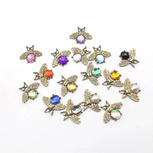 May include: A collection of decorative bee-shaped embellishments. Each bee features a gold-colored metal body and wings, with a central gemstone or pearl in various colors, including pink, purple, blue, green, and yellow. The embellishments are scattered on a white surface.