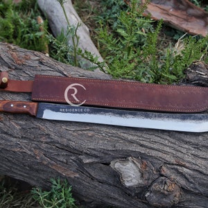 May include: A large, hand-forged machete with a wooden handle and a leather sheath. The blade is silver, and the handle is a dark brown. The sheath is brown leather. The text "RESIDENCE CO." is visible on the blade.