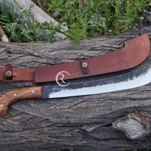 May include: A brown leather sheath with a decorative design and a metal button. A large, curved, silver blade with a wooden handle is partially inserted into the sheath. The blade has a dark, textured finish. The sheath and blade are lying on a rough, brown wooden surface.