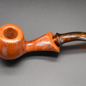 Puede incluir: Una pipa de madera con una cazoleta marrón y una boquilla marrón y ámbar.