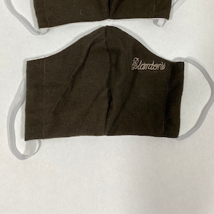 May include: Two brown face masks with white elastic ear loops. The masks have the word "Blanton's" embroidered in a gold color on the front. The masks are made of a soft, textured fabric.