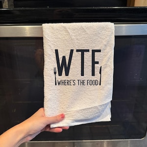 May include: White kitchen towel with the text "WTF WHERE'S THE FOOD" in black. A fork and knife are printed on either side of the text. The towel is hanging on an oven door.