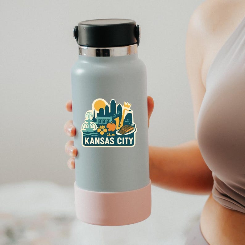 Puede incluir: Una botella de agua azul claro con tapa negra y base rosa. Una pegatina en la botella presenta un dise&ntilde;o del horizonte de Kansas City con un sol, edificios y el texto "KANSAS CITY".