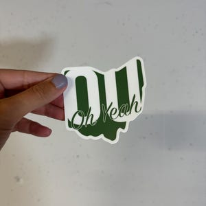 May include: A green and white sticker with the text "OU" and "Oh Yeah!" in a stylized font. The sticker is shaped like the state of Ohio.