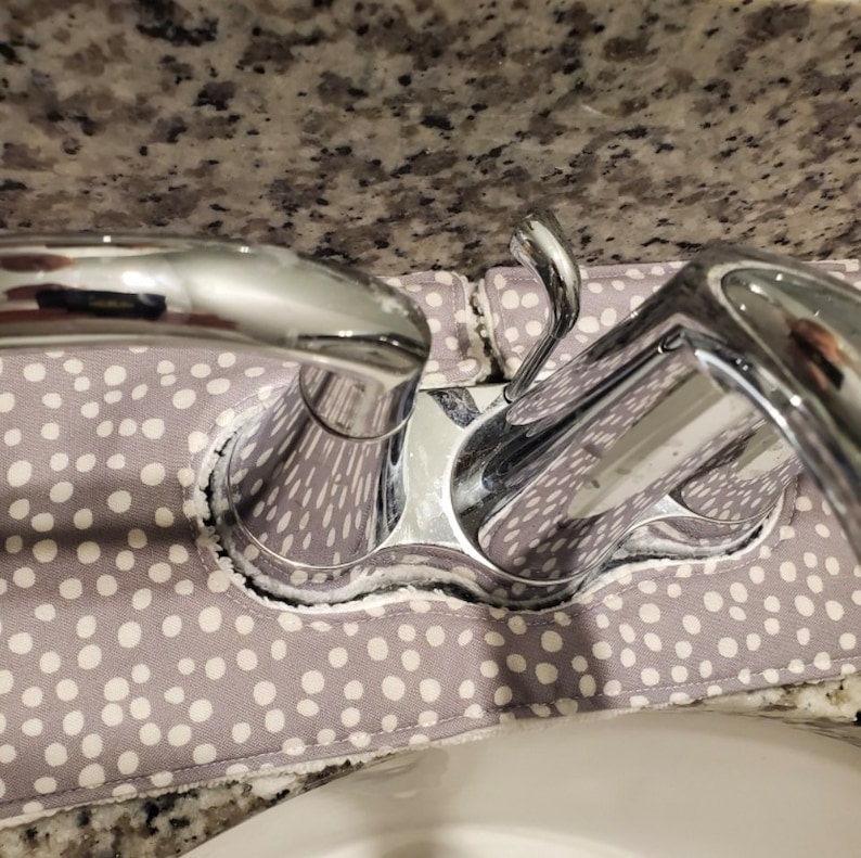 May include: A grey fabric faucet absorbent mat featuring a white polka dot pattern. The rectangular mat is positioned around the base of a chrome bathroom faucet, designed to catch water splashes and keep the surrounding countertop area dry.