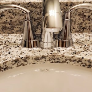 May include: A polished chrome bathroom tap with two curved handles and a central spout, mounted on a speckled beige and grey granite countertop above a white ceramic sink basin that features the word AVALON printed in small black letters.
