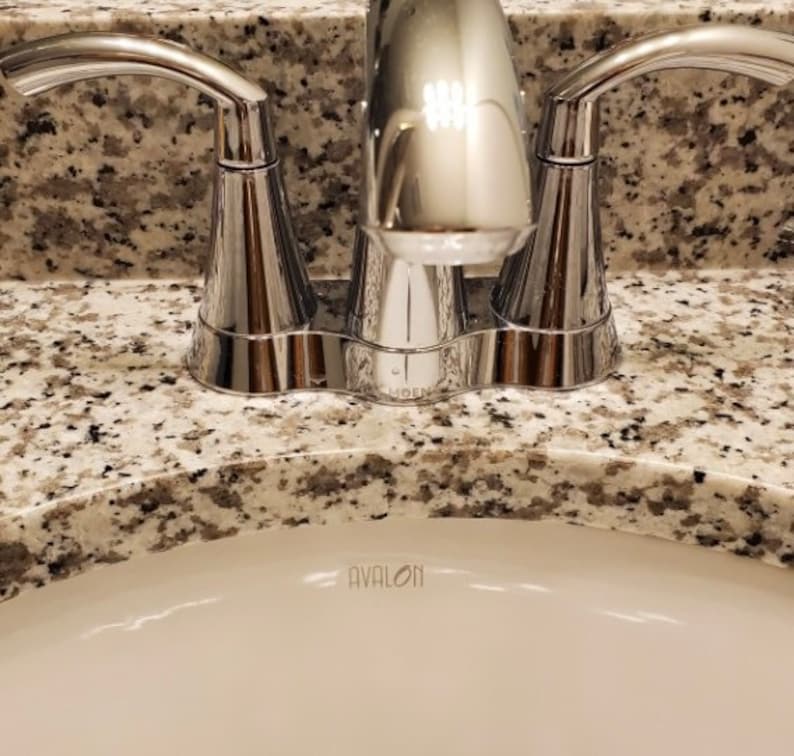 May include: A polished chrome bathroom tap with two curved handles and a central spout, mounted on a speckled beige and grey granite countertop above a white ceramic sink basin that features the word AVALON printed in small black letters.