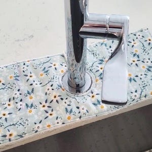 Op de afbeelding: Een absorberend pad voor een kraan met bloemenprint. Het pad is lichtblauw met een patroon van witte en gele bloemen en groene bladeren. Het pad is ontworpen om water te absorberen en het aanrecht te beschermen tegen waterschade.