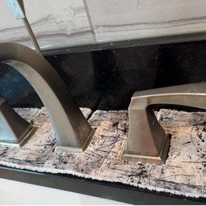 May include: A brushed nickel bathroom tap set with a high-arched spout and two lever handles. The tap base is surrounded by a rectangular, absorbent fabric mat featuring a distressed white and black textured pattern with dark scratch marks.