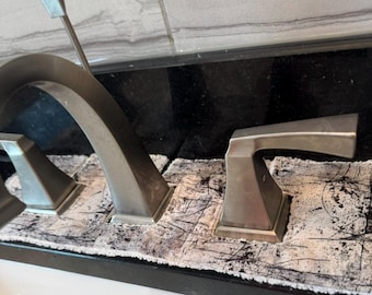 Op de afbeelding: Een badkamerkraan van geborsteld nikkel met een hoge booguitloop en twee hendels. De basis van de kraan is omgeven door een rechthoekige, absorberende stoffen mat met een gestructureerd wit en zwart patroon met donkere krasmarkeringen.