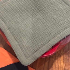 May include: A square, sage green quilted potholder rests on a red plate. The potholder has a textured, crinkled appearance and a stitched border. The corner of an orange and navy blue garment is visible in the lower portion of the image.