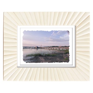 May include: A framed landscape photograph featuring a coastal scene with a pier extending into the water. The frame is a light cream color with a textured, fan-like design. The photograph captures a serene waterscape.