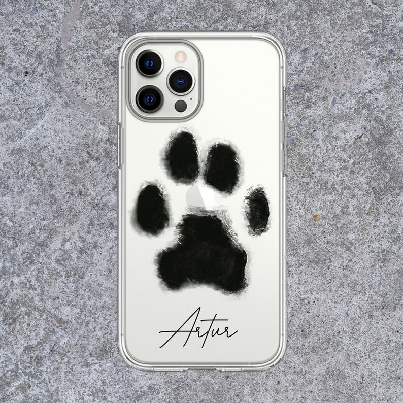 May include: Clear phone case featuring a large, black paw print design. The name "Artur" is written in a cursive font below the paw print. The case is shown on a gray, textured surface.