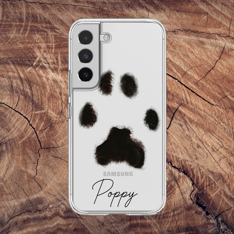 May include: Clear phone case with a black and white paw print design. The case is for a Samsung phone and has the name "Poppy" in a script font.