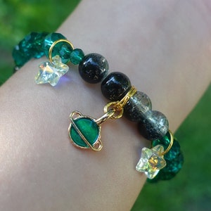 Peut inclure: Un bracelet composé de perles vertes et noires avec un pendentif en or représentant une planète avec des anneaux. Le bracelet est également orné de petits pendentifs en forme d'étoiles dorées.
