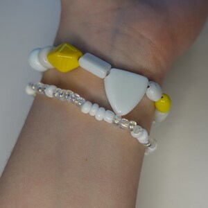 May include: Two bracelets on a wrist. The top bracelet has white and yellow beads, including a large white triangle bead. The bottom bracelet is made of small white beads.