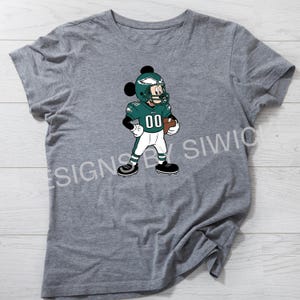 Puede incluir: Camiseta gris con un personaje de dibujos animados con uniforme de f&uacute;tbol americano. El personaje lleva un casco y una camiseta verdes con el n&uacute;mero "00" y sostiene un bal&oacute;n de f&uacute;tbol. El dise&ntilde;o est&aacute; centrado en la camiseta.
