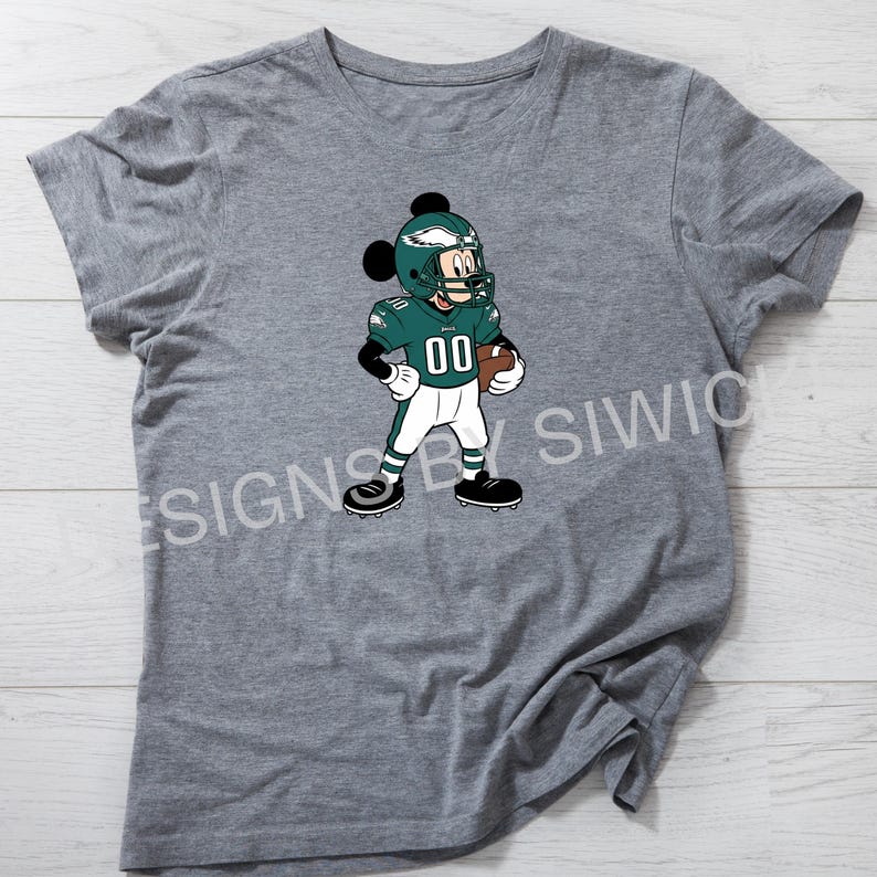 Puede incluir: Camiseta gris con un personaje de dibujos animados con uniforme de f&uacute;tbol americano. El personaje lleva un casco y una camiseta verdes con el n&uacute;mero "00" y sostiene un bal&oacute;n de f&uacute;tbol. El dise&ntilde;o est&aacute; centrado en la camiseta.