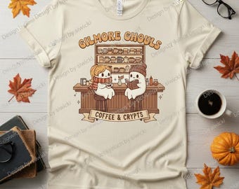 May include: Cream-colored t-shirt featuring a cartoon illustration with the text "GILMORE GHOULS" above a coffee shop scene. Two ghost characters are seated at the bar with the text "COFFEE & CRYPTS" below. Autumn leaves and a pumpkin are in the background.