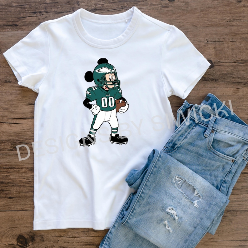 Puede incluir: Camiseta blanca con un personaje de dibujos animados con uniforme de f&uacute;tbol, n&uacute;mero 00. El personaje lleva un casco verde y sostiene un bal&oacute;n de f&uacute;tbol. La camiseta se presenta con unos vaqueros azules sobre una superficie de madera.