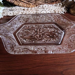 May include: Clear glass serving tray with an ornate floral and scroll design. The rectangular tray has a hexagonal center and handles on each end. The tray is approximately 12 inches long and 7 inches wide.