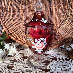 May include: A ruby red glass jar with a lid, featuring hand-painted white floral and rose designs. The jar sits on a clear, ornate base. The background includes a woven basket and a vintage lamp.