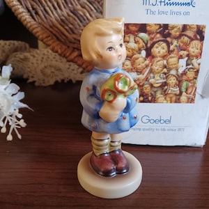 May include: A porcelain figurine of a young girl with blonde hair wearing a blue dress and holding a bouquet of green flowers. The figurine is standing on a round base and is part of a collection by Goebel.